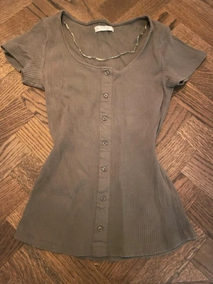 Ribbad grå topp med knappar - Snygg ribbad grå topp med korta ärmar och knappar längs hela framsidan. Toppen har en figurnära passform och en djup rundad halsringning. Perfekt att styla med jeans eller kjol.