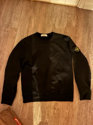 Svart sweatshirt från Stone Island - Säljer en svart sweatshirt från Stone Island med klassisk rund hals och ikonisk logotyp-patch på vänster ärm. Tröjan har ribbade muddar vid ärmslut och nederkant, perfekt för en clean och stilren look. Storlek S. Inga defekter. 