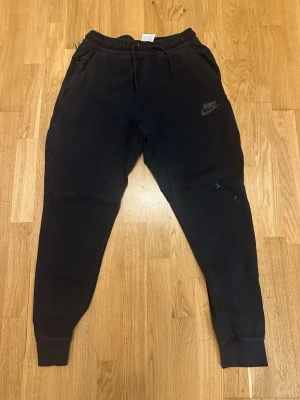 Svarta mjukisbyxor från Nike - Svarta mjukisbyxor från Nike med klassisk logga på vänster lår. Byxorna har snörning i midjan och muddar vid bensluten för en skön och avslappnad passform. Perfekta för chill eller träning. Tillverkade i mjukt bomullsmaterial. Paint splash obs!