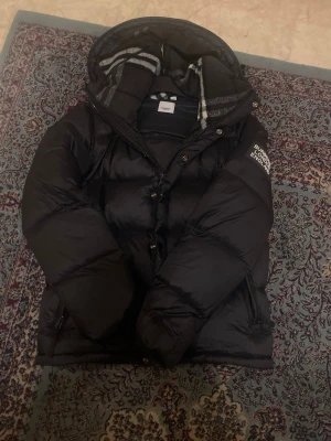 Svart dunjacka från Burberry - Snygg svart dunjacka från Burberry med klassiskt rutigt foder i huvan och tryck på ärmen. Helt oanvänd och perfekt för nuvarande vintern. Pris kan diskuteras