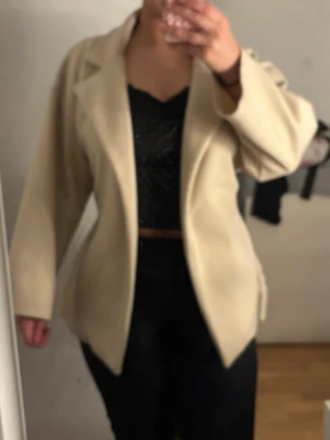 Beige kappa med bälte - Stilren beige kappa med bred krage och tillhörande bälte i midjan. Kappan har en klassisk, rak passform och är tillverkad i ett mjukt material som ger en snygg siluett. Perfekt för dig som vill ha en enkel men trendig look.