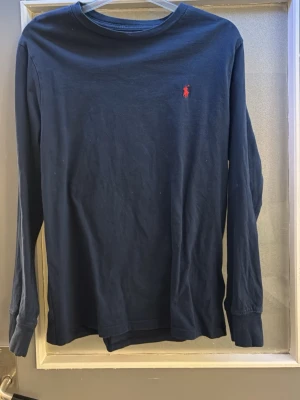 Marinblå långärmad topp från Polo Ralph Lauren - Klassisk marinblå långärmad topp från Polo Ralph Lauren med rund halsringning och den ikoniska röda logotypen broderad på bröstet. Tillverkad i mjuk bomull som känns skön mot huden. Perfekt att styla till jeans eller kjol. Obs osäker om den är äkta om det är avgörande för någon☺️ storleken är L/160 för en 14-16 åring