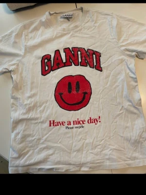 Vit Ganni t-shirt med smileytryck - Vit t-shirt från Ganni med stort rött smileytryck och texten 'Have a nice day! Please recycle' framtill. T-shirten har rund hals och korta ärmar. Perfekt för dig som gillar statement-plagg och coola tryck.