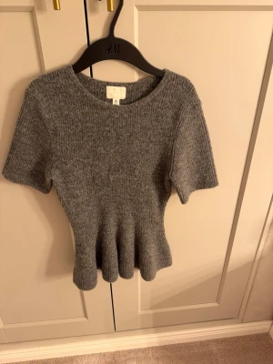 Grå ribbad stickad topp från H&M - Grå stickad topp från H&M med korta ärmar och ribbad struktur. Peplumtopp i mörkgrå. Använd få gånger vilket ger det bra skicket