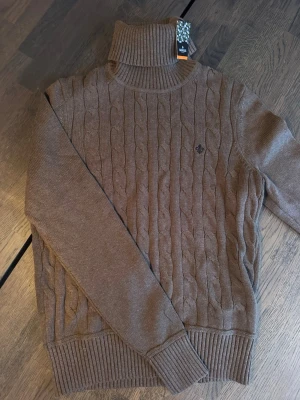 Stickad Morris tröja - Säljer en brun kabelstickad polotröja från Morris i storlek XS. Tröjan har hög krage, ribbade muddar och ett diskret Morris-emblem på bröstet. Perfekt för dig som gillar klassisk och stilren design med en mysig twist.