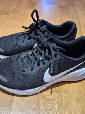 Svarta Nike sneakers - Snygga svarta Nike sneakers med vit sula. Skorna har rund tå, snörning och är tillverkade i ett lätt syntetmaterial som andas. Perfekta för dig som gillar sportig stil.