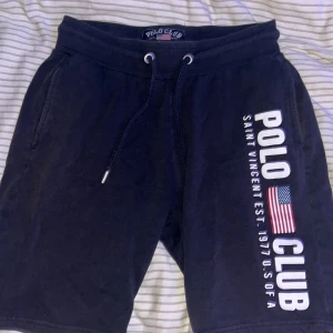Mörkblå shorts från Polo Club S - Mörkblå shorts från Polo Club med vit logga och amerikansk flagga tryckt på ena benet. Sköna med snörning i midjan, sidofickor och en bakficka. Perfekta för chill dagar eller träning. Tillverkade i mjuk bomull för extra komfort.