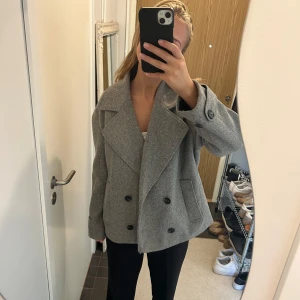 Grå skepparkavaj med stora slag - Snygg grå skepparkavaj med dubbelknäppning och stora slag. Jackan har två fickor framtill, stora svarta knappar och en loose passform. Perfekt för dig som gillar en stilren och trendig look.