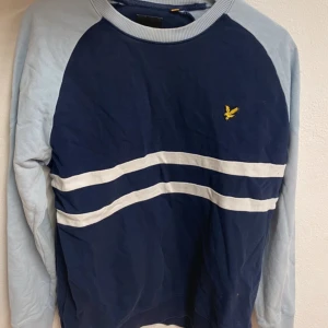 Blå sweatshirt från Lyle & Scott - Snygg blå sweatshirt från Lyle & Scott med ljusblå ärmar och vita ränder över bröstet. Tröjan har rund halsringning och det klassiska gula logobroderiet på bröstet. Perfekt för en chill och sportig stil.