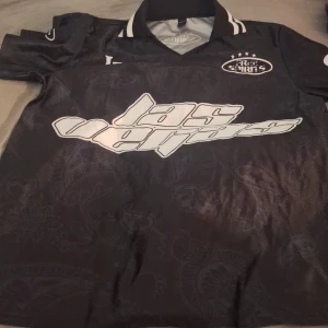 Svart fotbollströja Las Vegas Romwe XL - Cool svart fotbollströja från Romwe i storlek XL med vit text 'las vegas' på framsidan och 'Groovy 23' på ryggen. Tröjan har krage med vita detaljer och diskret paisleymönster i tyget. Perfekt för dig som vill sticka ut på planen eller gatan.