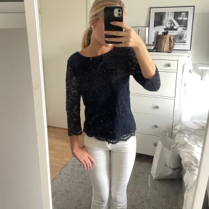 Mörkblå spetsblus med trekvartsärm - Säljer en mörkblå blus i spets med trekvartsärmar och rund halsringning. Blusen har fina spetsdetaljer och vågig kant nertill för en extra snygg touch. Perfekt att styla med jeans eller kjol för en trendig look.