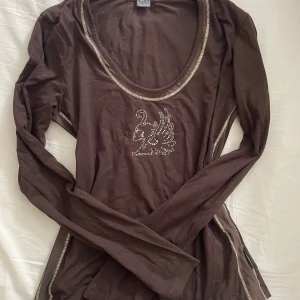 Långärmad topp med strass - Y2K Vintage Långärmad Topp CAPPOPERA Brun Strass Griffon - Made in Italy Djup chokladbrun färg som är enkel att matcha. Dekorerad med snygga guldfärgade kontrastsömmar runt halsen och längs sidorna. Motiv på svan med vingar gjort i glittrande strass/rhinestones.  #Y2K #vintage #2000s #retro #aesthetic #y2kfashion #rhinestones #strass #indie #CAPPOPERA #italien #madeinitaly #longsleeve #långärmad #topp #slimfit #bodycon #brun #chokladbrun #kontrastsöm #exposedseam #griffon