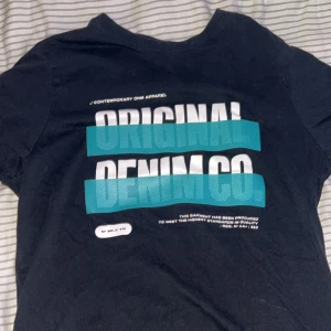 Svart t-shirt FSBN med tryck S - Svart t-shirt från FSBN i storlek S med stort tryck 'ORIGINAL DENIM CO.' i vitt och turkost framtill. Klassisk rund hals och korta ärmar. Regular fit-modell, perfekt för en avslappnad streetstil. Tillverkad i mjuk bomull. Har aldrig använt den och det ligger bara här hemma i onödan.