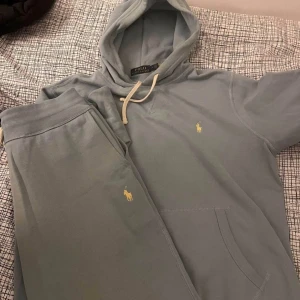 Ljusblå hoodie och byxor från Polo Ralph Lauren - Säljer en ljusblå hoodie från Polo Ralph Lauren med klassisk gul broderad logga på bröstet. Tröjan har huva med vita snören och en stor magficka. Mjuk bomullskvalitet som är perfekt för chill dagar. Passar dig som gillar stilren och enkel design.