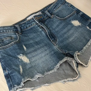 Blå jeansshorts med fransig kant - Snygga blå jeansshorts med slitna detaljer och fransig kant nertill. Klassisk femficksmodell med dragkedja och knapp framtill. Perfekta för sommaren och ger en avslappnad vibe 💙