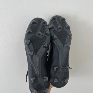 Svarta Nike fotbollsskor med rosa swoosh - Säljer ett par svarta Nike fotbollsskor med snygg rosa swoosh på sidan. Skorna har snörning, platt sula med dobbar och är tillverkade i syntetmaterial. Perfekta för dig som vill sticka ut på planen med stilrena och sportiga skor.