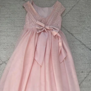 Finkänning med spets barn - Superfin ljusrosa långklänning från H&M med spets upptill och en stor rosett framtill i midjan. Klänningen har ett vackert fall, är ärmlös och har flera lager tyg i kjolen för extra volym. Perfekt för speciella tillfällen när man vill känna sig extra fin. Endast använd en gång, mycket bra skick.