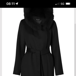 Svart kappa med fuskpäls från Hollies - Stilren svart kappa från Hollies med stor huva kantad av fuskpäls. Kappan har bälte i midjan för snygg passform och långa ärmar. Perfekt för kalla dagar när du vill vara både varm och snygg nypris 8000. Pris kan diskuteras vid snabbt köp