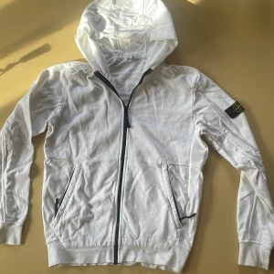 Vit hoodie från Stone Island Junior - Vit hoodie från Stone Island Junior med dragkedja framtill och klassisk logga på ärmen. Passar dig som är ca 170-180. Tröjan har huva, två fickor med dragkedja och långa ärmar. Tillverkad i mjuk bomull, perfekt för en clean och stilren look.