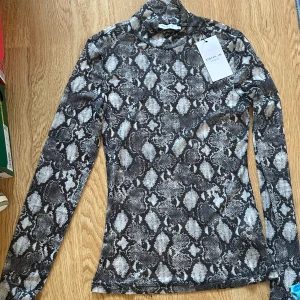 Polotopp med ormmönster från Stockh LM - Snygg polotopp från Stockh LM i svart, grått och vitt med coolt ormmönster. Topp med lång ärm och tight passform, perfekt för att ge outfiten edge. Mjukt och stretchigt material som sitter skönt mot kroppen.