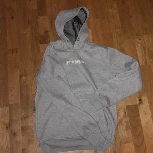 Grå Palm Angels hoodie med tryck - Snygg grå hoodie från Palm Angels med vit logga tryckt på både bröstet och ryggen. Klassisk känguruficka framtill, ribbade muddar och huva med meshfoder. Perfekt för dig som gillar streetwear och vill ha en stilren look.
