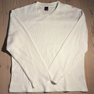 Vit långärmad tröja från derschütze - Vit långärmad tröja från derschütze i strukturerad bomull med broderad blomkvist i rosa och svart på axeln. Tröjan har rund halsringning och en clean, stilren look med snygg detalj på ryggen. Perfekt för dig som gillar unika broderier och enkel design.