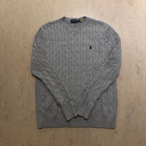 Stickat Ralph Lauren tröja grå - Säljer en klassisk grå kabelstickad tröja från Polo Ralph Lauren. Tröjan har rund halsringning, lång ärm och den ikoniska broderade loggan på bröstet. Perfekt för lager-på-lager och enkel att matcha med jeans eller chinos.