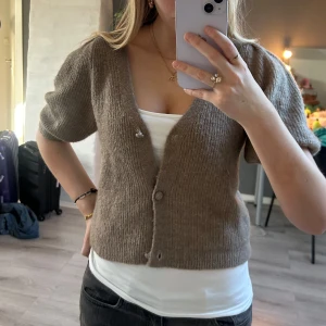 Brun stickad kofta  - Kort brun stickad kofta med v-ringning och knappar framtill. Koftan har korta ärmar och en ribbad struktur. Har små slitningar som syns på de sista bilderna men inget man tänker på!