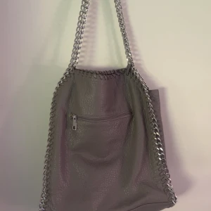 Stella McCartney väska - Säljer en ljus grå Stella McCartney liknande väska. Väskan har ett stort fack som stängs med dragkedja och tre innerfack varav ett av dom stängs med dragkedja. Väskan har även ett ytterfack. Väskan rymmer väldigt mycket och är perfekt till skolan då bland annat datorn får plats💞 väskan har en mindre defekt som syns på sista bilden, men som man inte märker av när väskan används. 