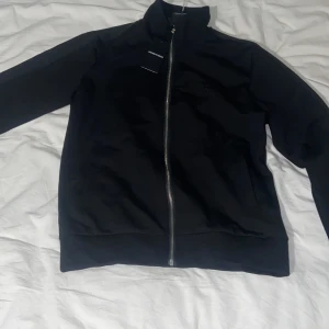 Svart bomberjacka från Emporio Armani - Svart bomberjacka från Emporio Armani med dragkedja framtill, hög krage och diskret logga på bröstet. Snygga detaljer längs axlarna med ton-i-ton text. Jackan är i ett mjukt material och har en clean, modern look.