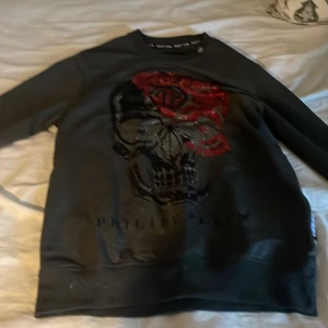 Svart tröja med döskalle från Philipp Plein - Cool svart tröja från Philipp Plein i 100% bomull med ett stort döskalletryck framtill i svart och rött. Tröjan har rund halsringning och långa ärmar. Perfekt statement piece för dig som vill sticka ut.