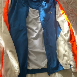 Blå och vit träningsdress från Lacoste - Säljer en snygg träningsdress från Lacoste i blått, vitt och orange. Jackan har dragkedja och elastiska muddar, byxorna har snörning i midjan och logga på benet. Materialet är lätt och andas, perfekt för sport eller chill. Klassisk krokodil på både jacka och byxa.