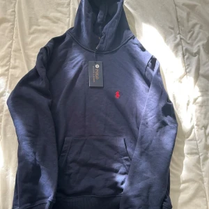 Mörkblå hoodie från Polo Ralph Lauren - Mörkblå hoodie från Polo Ralph Lauren med klassisk röd broderad logga på bröstet. Tröjan har huva, känguruficka och ribbade muddar. Perfekt för dig som gillar stilren och sportig look.