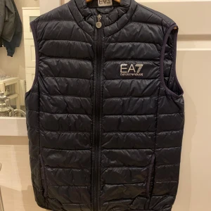 Mörkblå dunväst från Emporio Armani - Mörkblå dunväst från Emporio Armani EA7 med dragkedja framtill och logga på bröstet.  Knappt använd. Köptes från Zalando ny för 1200kr. Köparen står för frakt!
