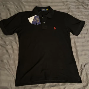 Svart pikétröja från Polo Ralph Lauren - Säljer en helt ny svart pikétröja från Polo Ralph Lauren i bomull med röd broderad logga på bröstet. Slim fit-modell som sitter snyggt och stilrent. Mitt pris 600kr