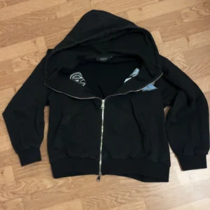 Svart Trapstar hoodie med dragkedja - Svart hoodie från Trapstar med stor huva och coolt broderat Trapstar-tryck i blått, vitt och svart över bröstet. Tröjan har hel dragkedja, ribbade muddar och två fickor framtill. Perfekt för dig som vill ha streetstyle-vibe. Passar mer som S. PRIS KAN DISKUTERAS.