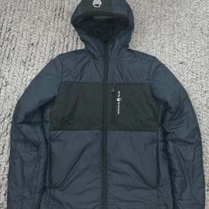 Svart och mörkblå pufferjacka Sail Racing - Pufferjacka från Sail Racing i svart och mörkblått med huva och dragkedja framtill. Jackan har en bröstficka med logga, två sidofickor med dragkedja och en quiltad look. Perfekt för kalla dagar och har en sportig vibe.