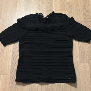 Svart blus med volang och spets - Svart blus med horisontella ribbade detaljer och volang längs bröstet. Blusen har spets vid halsringningen och halv-långa ärmar. Perfekt för dig som gillar en stilren men ändå detaljrik look. (Den är lite sliten där bak men går att fixas)