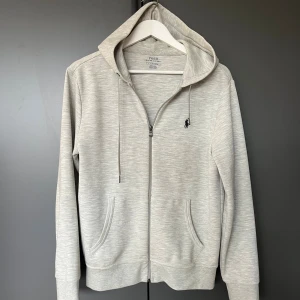 Ljusgrå hoodie från Polo Ralph Lauren - Säljer en stilren ljusgrå hoodie från Polo Ralph Lauren med dragkedja, huva och snörning. Tröjan har två fickor framtill och en liten broderad logga på bröstet. Perfekt för en chill och avslappnad look.