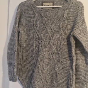 Grå stickad tröja från Bondelid - Mysig grå stickad tröja från Bondelid med flätstickat mönster framtill och broderad detalj på ärmen. Tröjan har rund halsringning och långa ärmar, perfekt för kyliga dagar. Snygg och enkel att matcha med jeans eller kjol.