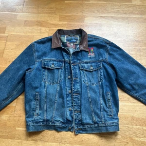 Vintage jeansjacka Upstream XL Kellogg’s corn flakes - Retro jeansjacka från Upstream med broderad Kellogg's Corn Flakes-logga på bröstet. Jackan har klassisk blå denim, brunt skinnkrage och rutigt innerfoder. Två bröstfickor med lock och knappar samt sidofickor. Perfekt för dig som gillar unik streetstyle.