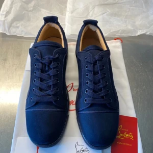 Mörkblå sneakers från Christian Louboutin - Snygga mörkblå sneakers från Christian Louboutin i mocka med klassisk röd sula. Skorna har rund tå, platt sula och snörning framtill. Diskret broderad logga baktill och stilren design som sticker ut med den ikoniska röda undersidan.