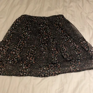 Blommig svart kortkjol med volang - Supersöt svart kortkjol med småblommigt mönster i rosa, vitt och blått. Kjolen har volangdetaljer och resår i midjan för en bekväm passform. Perfekt för dig som gillar en romantisk och trendig stil.