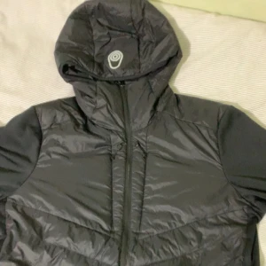 Svart pufferjacka med huva - Svart pufferjacka med huva och dragkedja framtill. Jackan har en glansig yta och quiltade partier på bröst och mage. Perfekt för kyliga dagar och har en sportig vibe. Reflekterande detalj på huvan.