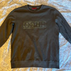 Svart sweatshirt från Hugo Boss - Säljer en svart sweatshirt från Hugo Boss med diskret logga framtill. Tröjan har rund halsringning och långa ärmar, perfekt för en clean och stilren look. Tillverkad i mjuk bomull som känns skön mot huden.
