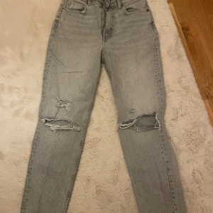 Grå ripped jeans från Zara - Snygga ljusgrå jeans från Zara i storlek 34 med slitna detaljer och hål över knäna. Hög midja och klassisk femficksmodell. Perfekta för en avslappnad och trendig look. Tillverkade i bomull för skön passform. Byxornas längd är 96 cm. 