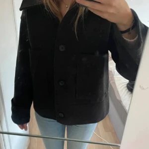 Svart höstkappa med stora fickor - Säljer en svart/grå kappa med stora framfickor och klassisk krage. Jackan har knäppning med stora knappar framtill och raka ärmar. Perfekt för lager-på-lager och enkel att styla till jeans eller kjol. Köptes för 900kr🌸