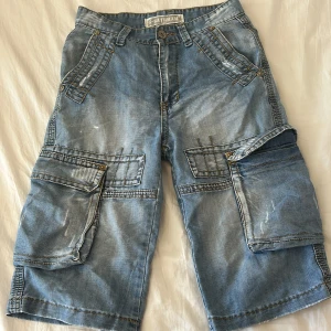 Blå jeansshorts med cargofickor G-Satrram - Säljer ett par blå jeansshorts från G-Satrram i storlek 29. De har flera stora cargofickor med lock och coola sömmar, samt bälteshällor. Shortsen är loose fit och har en klassisk midwaist. Perfekt för dig som gillar streetstyle och funktionella detaljer.