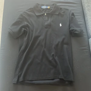 Svart Polo Ralph Lauren t-shirt - Svart t-shirt från Polo Ralph Lauren med klassisk pikékrage och två knappar framtill. Ikonisk vit broderad logga på bröstet. Modellen är kortärmad och har en normal passform. Perfekt för dig som gillar stilrena och tidlösa plagg.