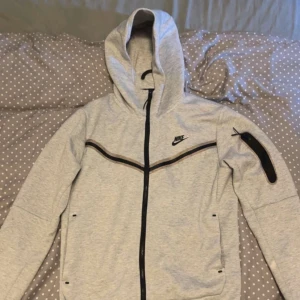 Grå Nike hoodie med dragkedja - Snygg ljusgrå hoodie från Nike med svart dragkedja och svarta detaljer. Tröjan har huva, lång ärm och en extra ficka med dragkedja på ena ärmen. Perfekt för en sportig och avslappnad stil.